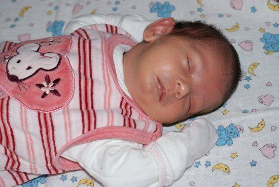 Ein Babymädchen schläft auf einem Bett mit einem pink-weiß gestreiften Strampler und einem rosa Lätzchen mit einem Cartoon-Charakter darauf.