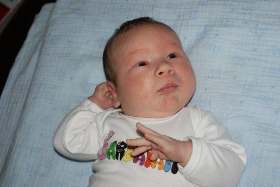 Ein Baby liegt auf einer blauen Decke und trägt ein weißes Langarmshirt mit 'SARAHAN' in bunten Buchstaben. Das Baby hat einen leicht geöffneten Mund und schaut zur Seite.
