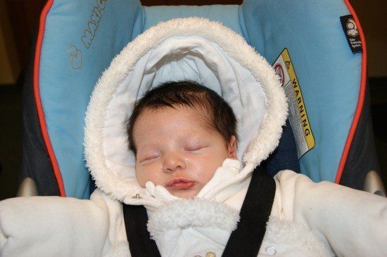 Ein Baby schläft in einem Autositz und trägt einen weißen Wintermantel mit Kapuze. Der Autositz ist blau mit orangefarbenem Rand.