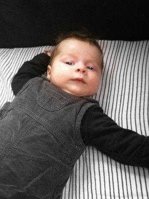 Ein Baby liegt auf einer gestreiften Bettdecke, trägt einen grauen Overall und hat einen ernsten Gesichtsausdruck mit den Händen auf dem Bett.