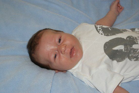Ein Baby liegt auf einer blauen Decke und trägt ein weißes T-Shirt mit einem grauen Grafikdesign. Das Baby hat braune Haare und schaut direkt in die Kamera.