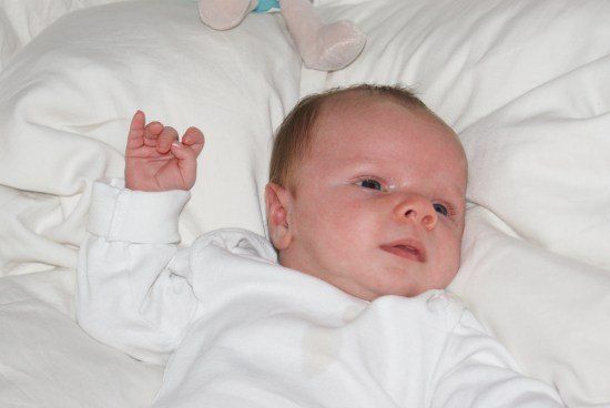 Ein Baby liegt auf einer weißen Decke, trägt ein weißes Strampler und schaut mit neugierigem Blick direkt in die Kamera.
