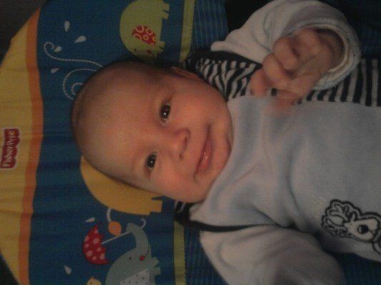 Ein lächelndes Baby liegt auf einer bunten Spielmatte mit Comicfiguren und trägt ein weißes Shirt mit einem Hirschdesign.
