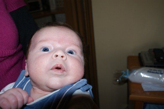 Ein Baby mit blauen Augen und einem gestreiften blauen und weißen Tuch wird von einer Person in einem lila Pullover gehalten.