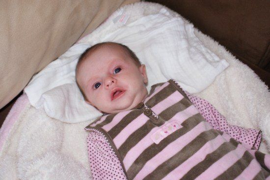Ein Babymädchen liegt auf einer weißen Decke und trägt ein pink-braun gestreiftes Outfit. Sie hat einen glücklichen Ausdruck auf dem Gesicht.