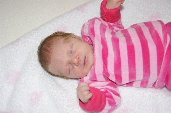 Ein Babymädchen schläft auf einer weißen Decke und trägt rosa-weiß gestreifte Schlafanzug. Ihre Augen sind geschlossen, und sie hat ein kleines Lächeln im Gesicht.