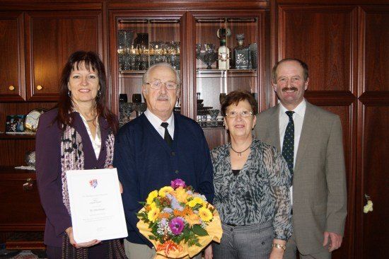 Vier Personen stehen in einem Raum mit einem hölzernen Schrank dahinter. Eine Frau hält ein Zertifikat und einen Blumenstrauß. Ein Mann neben ihr trägt eine Krawatte und eine Brille. Eine weitere Frau neben ihm trägt eine Brille und eine Halskette. Ein Mann auf der rechten Seite trägt einen Anzug und eine Krawatte.