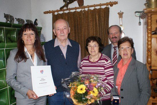 Eine Gruppe von Menschen steht in einem Raum, ein Mann hält ein Zertifikat und einen Blumenstrauß. Eine Frau links trägt einen grauen Anzug, und eine Frau rechts hält eine Medaille. Hinter ihnen ist eine Wand mit einer Uhr und einem Vorhang.