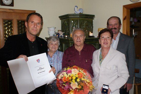 Eine Gruppe von Menschen steht in einem Raum, ein Mann hält ein Zertifikat und einen Blumenstrauß und eine Frau hält eine Medaille.
