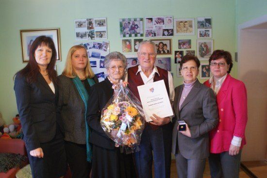 Eine Gruppe von Menschen steht in einem Raum mit einer grünen Wand, hält einen Blumenstrauß und ein Zertifikat. Eine ältere Frau und ein Mann stehen in der Mitte.