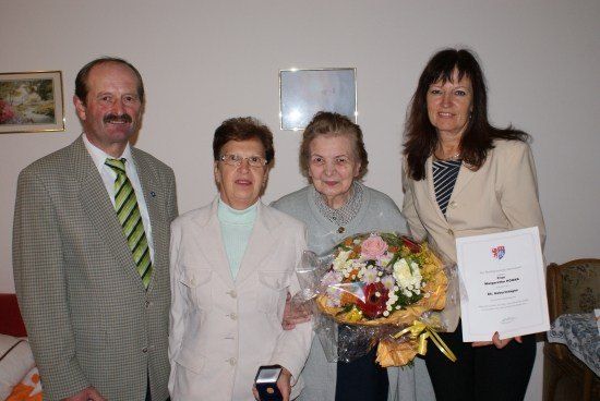 Vier Menschen stehen in einem Raum, zwei Frauen halten einen Blumenstrauß und ein Zertifikat, während der Mann links lächelt.