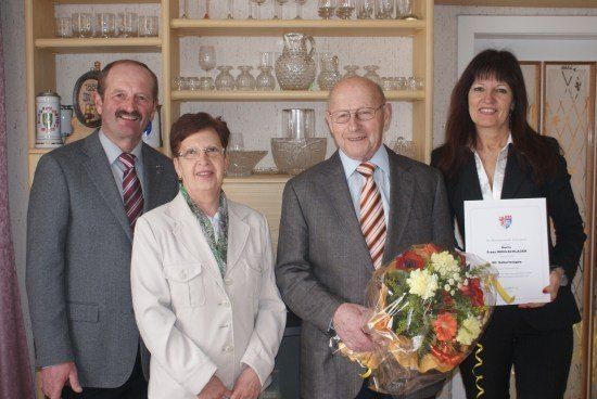 Vier Personen, zwei Männer und zwei Frauen, stehen zusammen und halten einen Blumenstrauß und ein Zertifikat. Sie sind formell gekleidet und scheinen eine Leistung zu feiern. Hinter ihnen steht ein Holzregal, das mit Glaswaren gefüllt ist.