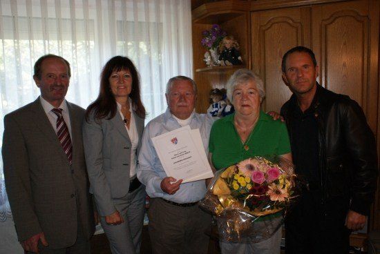 Fünf Personen stehen lächelnd in einem Raum. Der Mann in der Mitte hält ein Zertifikat und einen Blumenstrauß. Hinter ihnen steht ein Holzschrank mit einer Puppe und Blumen.