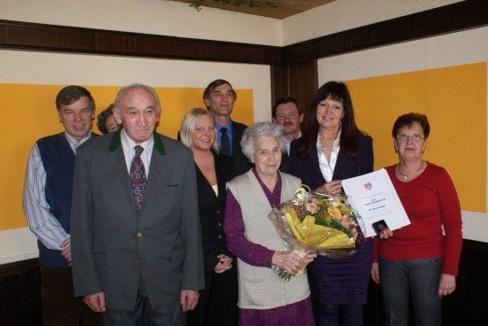Eine Gruppe von Menschen, darunter eine ältere Frau mit Blumen, steht in einem Raum. Sie alle lächeln und scheinen ein Ereignis zu feiern. Die Frau hält einen Blumenstrauß und ein Zertifikat.
