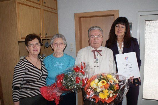 Vier Personen stehen in einem Raum, zwei Frauen halten Blumensträuße und ein Mann ein Zertifikat. Der Mann trägt eine Schleife und die Frau rechts hält ein Zertifikat.
