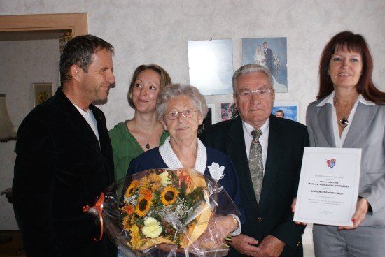 Eine Gruppe von Menschen, einschließlich einer älteren Frau, die einen Blumenstrauß hält, stehen zusammen und lächeln für ein Foto. Die ältere Frau hält eine Urkunde.