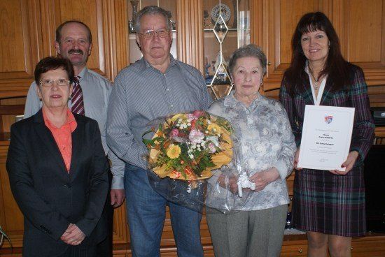 Fünf Personen stehen in einem Raum, zwei Frauen halten einen Blumenstrauß und ein Zertifikat, und drei Männer stehen hinter ihnen. Die Frau auf der rechten Seite lächelt und hält ein Zertifikat. Die Frau auf der linken Seite hält einen Blumenstrauß. Der Mann in der Mitte trägt eine Brille. Hinter ihnen steht ein hölzerner Schrank mit Glastüren.