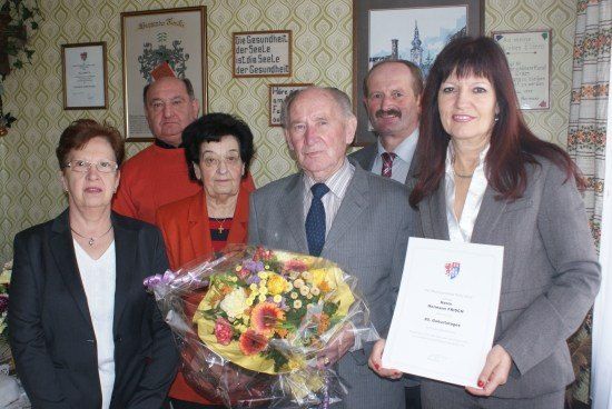 Eine Gruppe von Menschen steht lächelnd zusammen, einer hält einen Blumenstrauß und der andere ein Zertifikat. Sie befinden sich in einem Raum mit dekorativer Tapete.