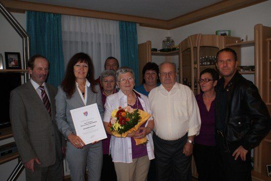 Eine Gruppe von Menschen steht in einem Raum, lächelt und posiert für ein Foto. Eine Frau in der Mitte hält ein Zertifikat und einen Blumenstrauß. Dahinter befinden sich Regale mit verschiedenen Gegenständen.