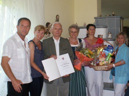 Eine Gruppe von Menschen steht zusammen und lächelt, einer hält ein Zertifikat und der andere einen Geschenkkorb. Hinter ihnen steht ein Kamin mit einer dekorativen Fliesengestaltung.