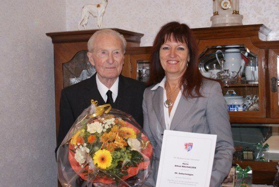 Ein älterer Mann und eine Frau stehen zusammen und halten einen Blumenstrauß und ein Zertifikat. Der Mann trägt einen Anzug und eine Krawatte, während die Frau in einem grauen Blazer ist. Hinter ihnen steht ein Holzschrank, gefüllt mit verschiedenen Gegenständen.