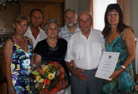 Eine Gruppe von Menschen posiert für ein Foto. Ein älterer Mann hält ein Zertifikat und eine Frau hält einen Blumenstrauß. Sie lächeln und scheinen zu feiern.