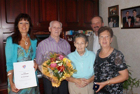 Fünf Personen stehen in einem Raum mit einem Blumenstrauß, einem Zertifikat und einer Trophäe. Die Frau auf der linken Seite hält ein Zertifikat und einen Blumenstrauß. Die Frau neben ihr hält eine Trophäe. Die Frau in der Mitte hält einen Blumenstrauß. Der Mann auf der rechten Seite trägt einen Anzug.