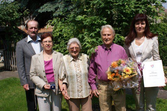 Fünf Personen stehen in einem Garten, lächeln und halten Blumen und ein Zertifikat. Die Frau in der Mitte trägt eine Brille und hält eine kleine blaue Schachtel. Der Mann rechts hält einen Blumenstrauß.