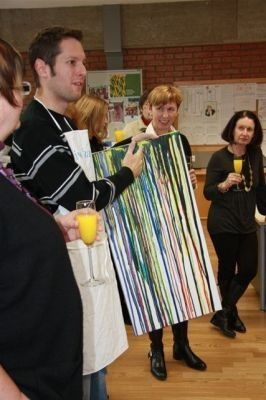 Eine Gruppe von Menschen steht um ein großes, farbenfrohes Gemälde herum. Ein Mann hält das Gemälde, während andere zusehen und Orangensaft trinken.