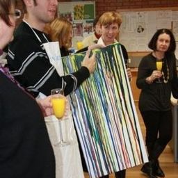 Eine Gruppe von Menschen steht um ein großes, farbenfrohes Gemälde herum. Ein Mann hält das Gemälde, während andere zusehen und Orangensaft trinken.