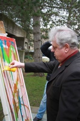 Ein älterer Mann malt ein farbenfrohes abstraktes Bild auf eine große Leinwand im Freien. Eine andere Person steht hinter ihm, teilweise verdeckt.
