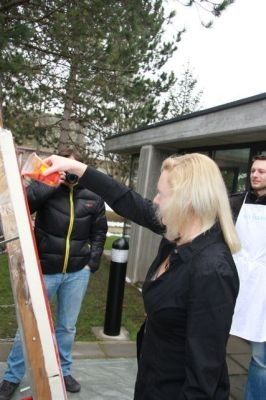 Eine Frau mit blonden Haaren malt draußen auf einer Leinwand, ein Mann in einer Jacke und Jeans steht hinter ihr. Ein weiterer Mann in einer weißen Schürze steht rechts von ihr.