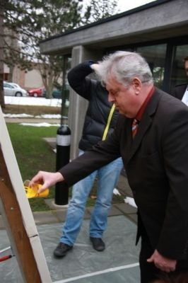Ein älterer Mann in einem Anzug gießt gelbe Flüssigkeit in einen Behälter, während ein anderer Mann in einer Jacke zusieht. Sie sind draußen in der Nähe eines Gebäudes.