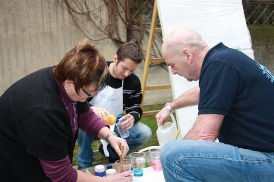 Drei Personen sind in einer praktischen Aktivität verwickelt, wobei eine Frau eine gelbe Flüssigkeit in einen Becher gießt, ein Mann in einer weißen Schürze beobachtet und ein anderer Mann in einem blauen T-Shirt eine rosa Flüssigkeit in einen Becher gießt.
