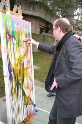 Ein Mann in einem schwarzen Anzug malt ein farbenfrohes abstraktes Werk auf einer großen Leinwand im Freien.