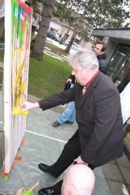 Ein älterer Mann in einem Anzug malt ein farbenfrohes abstraktes Design auf eine große Leinwand im Freien.