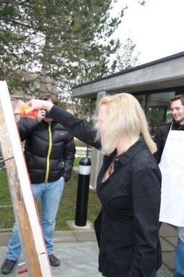 Eine Frau mit blonden Haaren malt draußen auf einer Leinwand, während ein Mann in einem schwarzen Mantel zuschaut. Ein anderer Mann in einer weißen Schürze steht in der Nähe.