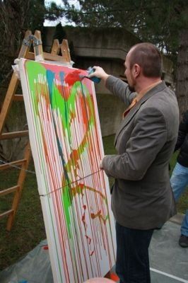 Ein Mann in einem grauen Anzug malt im Freien eine große Leinwand mit leuchtenden Farben.