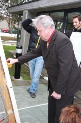 Ein älterer Mann in einem Anzug malt eine Holztafel, während zwei andere zusehen. Der Ort ist im Freien mit einem Gebäude im Hintergrund.