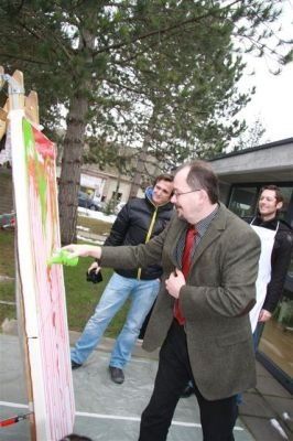 Ein Mann in Anzug und Krawatte malt draußen auf einer Leinwand, zwei Männer beobachten ihn dahinter.