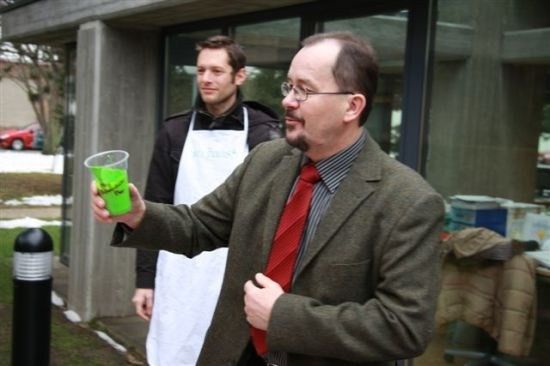 Ein Mann in Anzug und Krawatte hält einen grünen Drink vor einem Gebäude, ein anderer Mann in einer weißen Schürze steht dahinter.