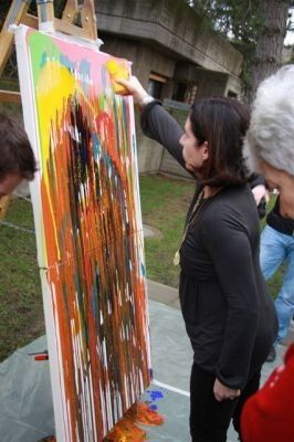 Eine Frau malt ein großes, abstraktes Bild mit kräftigen Farben. Andere schauen zu.