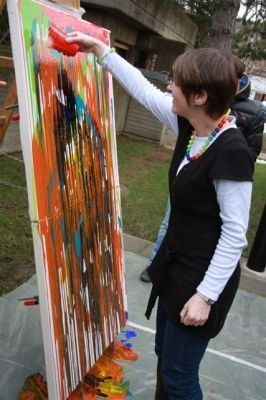 Eine Frau mit einem schwarzen Weste und einer Halskette malt eine große abstrakte Leinwand mit leuchtenden orangefarbenen und blauen Farben. Sie steht vor einem Gebäude mit einem Baum im Hintergrund.
