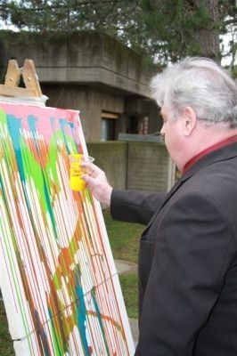 Ein älterer Mann in einem Anzug malt auf einer großen Leinwand mit leuchtenden Farben und hält einen gelben Becher in der Hand.