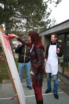Eine Frau in einem karierten Mantel und roten Hosen malt mit roter Farbe auf eine weiße Leinwand, während zwei Männer in Jacken und Jeans zusehen.