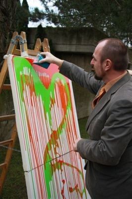 Ein Mann in einem grauen Anzug malt im Freien eine große Leinwand mit grüner und orangefarbener Farbe.