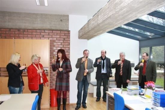 Eine Gruppe von Menschen in formeller Kleidung versammelt sich drinnen und hält Weingläser in der Hand und prostet. Der Raum hat eine Ziegelwand, einen Holzboden und eine Glasdecke.
