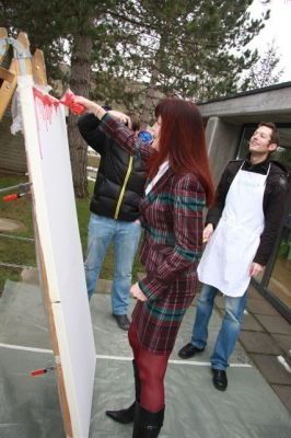 Eine Frau in einem karierten Mantel malt eine weiße Leinwand mit roter Farbe, während zwei Männer in Jacken und Schürzen hinter ihr stehen. Die Szene spielt im Freien mit Bäumen und einem Gebäude im Hintergrund.