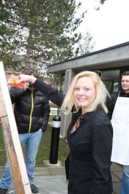 Eine Frau mit blonden Haaren hält eine Leiter und lächelt, während ein Mann hinter ihr ein Foto macht. Ein anderer Mann steht in der Nähe und trägt eine weiße Schürze.