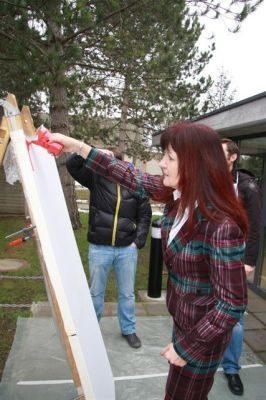 Eine Frau mit roten Haaren malt mit einem roten Marker auf eine weiße Tafel, während sie auf einer Betonplattform steht. Hinter ihr stehen zwei Männer, einer trägt eine Jacke und der andere Jeans. Bäume und ein Gebäude sind im Hintergrund.